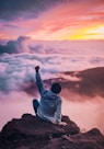 man sitting on mountain cliff facing white clouds rising one hand at golden hour