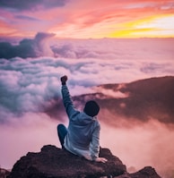 man sitting on mountain cliff facing white clouds rising one hand at golden hour