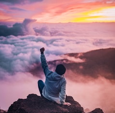 man sitting on mountain cliff facing white clouds rising one hand at golden hour