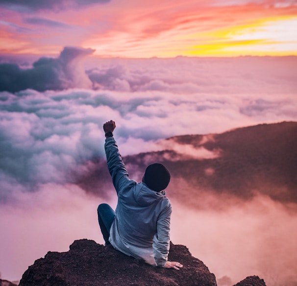 man sitting on mountain cliff facing white clouds rising one hand at golden hour