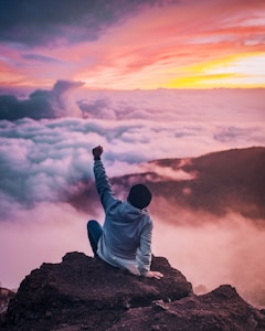man sitting on mountain cliff facing white clouds rising one hand at golden hour