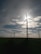 Several wind turbines are positioned in a vast open field, with the sun partially obscured by one of the turbine blades. The sky is dotted with scattered clouds, allowing beams of sunlight to break through. The landscape appears serene and expansive, with the focus on renewable energy sources.