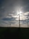 Several wind turbines are positioned in a vast open field, with the sun partially obscured by one of the turbine blades. The sky is dotted with scattered clouds, allowing beams of sunlight to break through. The landscape appears serene and expansive, with the focus on renewable energy sources.