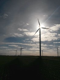 Several wind turbines are positioned in a vast open field, with the sun partially obscured by one of the turbine blades. The sky is dotted with scattered clouds, allowing beams of sunlight to break through. The landscape appears serene and expansive, with the focus on renewable energy sources.