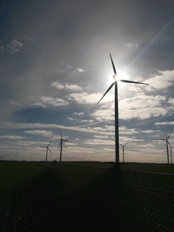 Several wind turbines are positioned in a vast open field, with the sun partially obscured by one of the turbine blades. The sky is dotted with scattered clouds, allowing beams of sunlight to break through. The landscape appears serene and expansive, with the focus on renewable energy sources.