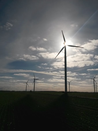 Several wind turbines are positioned in a vast open field, with the sun partially obscured by one of the turbine blades. The sky is dotted with scattered clouds, allowing beams of sunlight to break through. The landscape appears serene and expansive, with the focus on renewable energy sources.