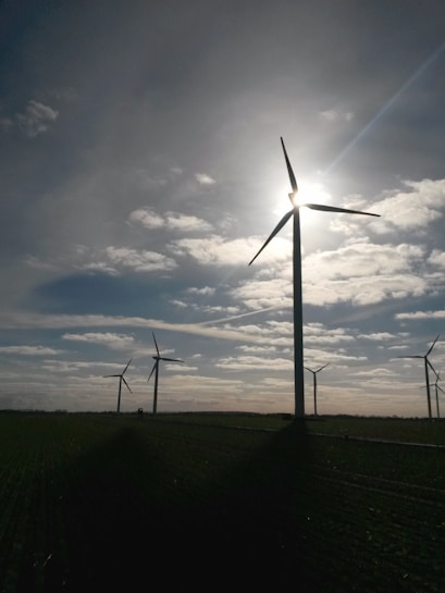 Several wind turbines are positioned in a vast open field, with the sun partially obscured by one of the turbine blades. The sky is dotted with scattered clouds, allowing beams of sunlight to break through. The landscape appears serene and expansive, with the focus on renewable energy sources.