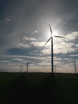 Several wind turbines are positioned in a vast open field, with the sun partially obscured by one of the turbine blades. The sky is dotted with scattered clouds, allowing beams of sunlight to break through. The landscape appears serene and expansive, with the focus on renewable energy sources.