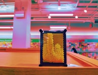 A pin art toy with yellow pins is placed on a wooden surface in a brightly colored room. The background is a blur of pink and teal hues, suggesting a retail setting like a store with shelves visible.