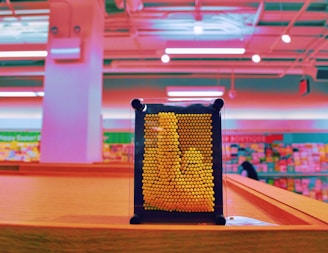 A pin art toy with yellow pins is placed on a wooden surface in a brightly colored room. The background is a blur of pink and teal hues, suggesting a retail setting like a store with shelves visible.