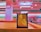 A pin art toy with yellow pins is placed on a wooden surface in a brightly colored room. The background is a blur of pink and teal hues, suggesting a retail setting like a store with shelves visible.