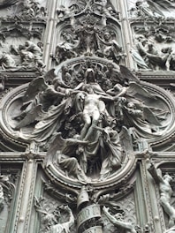 An intricate bronze relief sculpture depicting religious figures surrounded by ornate details and symbolic imagery. The central figure appears to be a robed individual with outstretched arms, framed by multiple figures, possibly angels, engaged in dynamic poses.