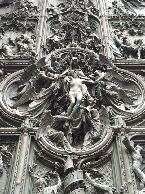 An intricate bronze relief sculpture depicting religious figures surrounded by ornate details and symbolic imagery. The central figure appears to be a robed individual with outstretched arms, framed by multiple figures, possibly angels, engaged in dynamic poses.