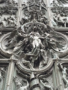 An intricate bronze relief sculpture depicting religious figures surrounded by ornate details and symbolic imagery. The central figure appears to be a robed individual with outstretched arms, framed by multiple figures, possibly angels, engaged in dynamic poses.