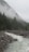 A serene mountain landscape featuring a flowing river flanked by rocky banks. Tall pine trees and dense forest cover the mountainsides, with a layer of mist hovering near the peaks, creating a mystical atmosphere.