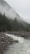 A serene mountain landscape featuring a flowing river flanked by rocky banks. Tall pine trees and dense forest cover the mountainsides, with a layer of mist hovering near the peaks, creating a mystical atmosphere.