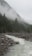 A serene mountain landscape featuring a flowing river flanked by rocky banks. Tall pine trees and dense forest cover the mountainsides, with a layer of mist hovering near the peaks, creating a mystical atmosphere.