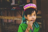 A child wearing traditional attire, adorned with a vibrant headdress featuring pink and yellow flowers. The child has brass neck rings and wears colorful garments with a green shawl. Decorative face paint is present. The background includes market stalls with various items.