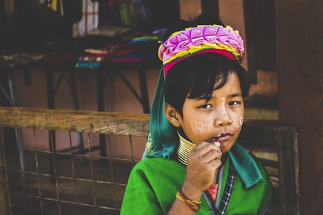 A child wearing traditional attire, adorned with a vibrant headdress featuring pink and yellow flowers. The child has brass neck rings and wears colorful garments with a green shawl. Decorative face paint is present. The background includes market stalls with various items.