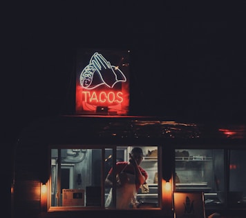 A dimly lit taco food truck with a brightly illuminated neon sign displaying the word 'TACOS' alongside a visual of a taco. Inside, a person is wearing a red shirt and apron, busily preparing or serving food. The surrounding environment is dark, highlighting the focused light from the truck and sign.