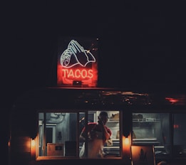 A dimly lit taco food truck with a brightly illuminated neon sign displaying the word 'TACOS' alongside a visual of a taco. Inside, a person is wearing a red shirt and apron, busily preparing or serving food. The surrounding environment is dark, highlighting the focused light from the truck and sign.