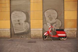 A red scooter is parked in front of a building with yellow walls. The building has two metal shutters painted with graffiti of large white letters. The ground is paved with gray cobblestones.