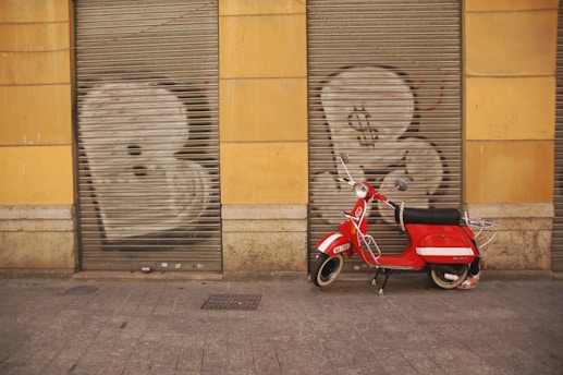 A red scooter is parked in front of a building with yellow walls. The building has two metal shutters painted with graffiti of large white letters. The ground is paved with gray cobblestones.
