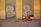 A red scooter is parked in front of a building with yellow walls. The building has two metal shutters painted with graffiti of large white letters. The ground is paved with gray cobblestones.