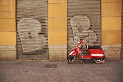 A red scooter is parked in front of a building with yellow walls. The building has two metal shutters painted with graffiti of large white letters. The ground is paved with gray cobblestones.