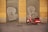 A red scooter is parked in front of a building with yellow walls. The building has two metal shutters painted with graffiti of large white letters. The ground is paved with gray cobblestones.