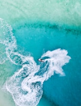 Aerial view of a person riding a jet ski across a vibrant turquoise ocean with trails of white frothy water following behind, creating dynamic patterns against the clear water.