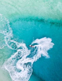 Aerial view of a person riding a jet ski across a vibrant turquoise ocean with trails of white frothy water following behind, creating dynamic patterns against the clear water.