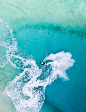 Aerial view of a person riding a jet ski across a vibrant turquoise ocean with trails of white frothy water following behind, creating dynamic patterns against the clear water.