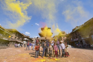 Children playing with colorful powders in an open square surrounded by traditional buildings, creating a festive and lively atmosphere. The sky is clear with a few white clouds, adding brightness to the scene.