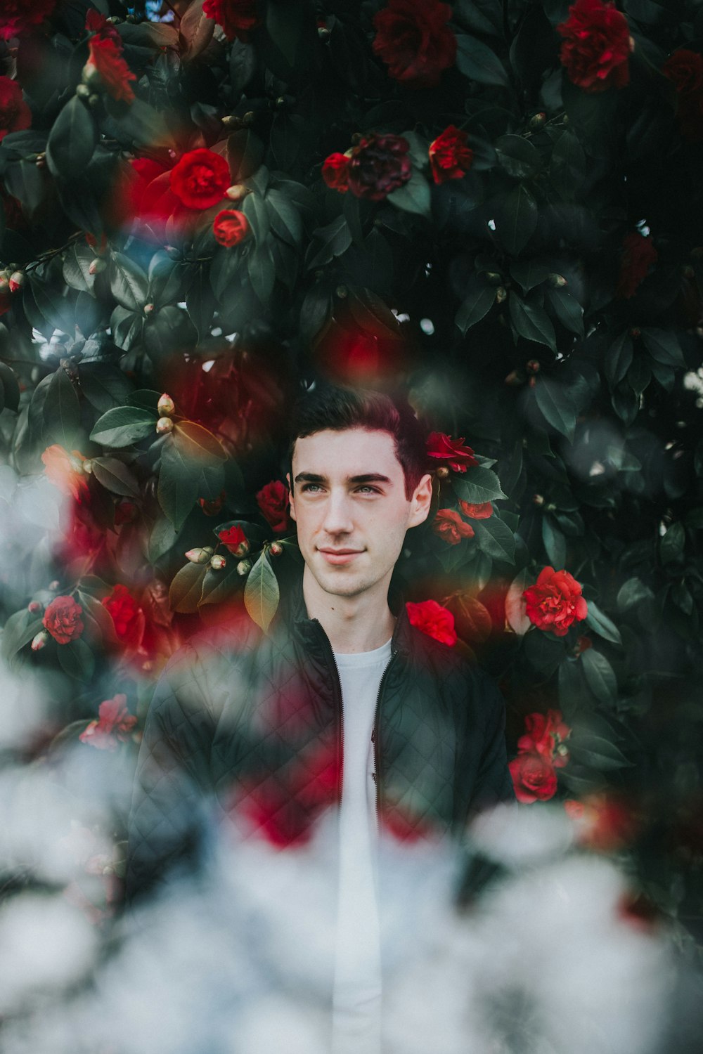 man in black zip-up jacket standing in frond of green plants and red flowers