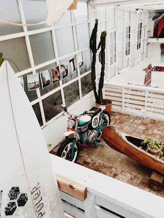 A cozy, rustic indoor setting with a vintage motorcycle marked 'Camp Vibes' and a carved wooden canoe filled with plants. Several photographs are displayed against a white grid-style window, and a surfboard is leaning against a wall. The area is surrounded by white paneling and large glass windows, with decorative rugs adding a touch of color.