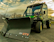 A rugged utility vehicle equipped with a large snow plow attachment. It is parked on a paved surface under a partly cloudy sky. The vehicle is mostly dark green and black, with prominent yellow wheels, and has a distinctive, sturdy construction suitable for heavy-duty tasks.