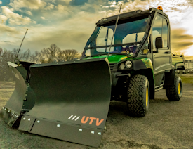 A rugged utility vehicle equipped with a large snow plow attachment. It is parked on a paved surface under a partly cloudy sky. The vehicle is mostly dark green and black, with prominent yellow wheels, and has a distinctive, sturdy construction suitable for heavy-duty tasks.