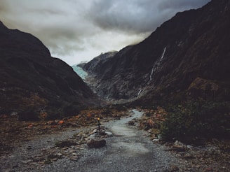 A rocky pathway leads through a dramatic landscape featuring steep, dark mountains on either side. Waterfalls cascade down the mountains, and in the distance, a glacier is visible beneath a cloudy sky.