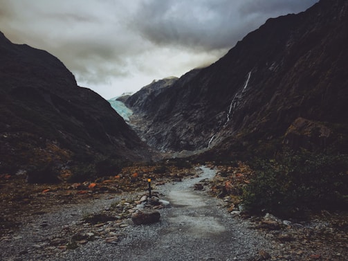 A rocky pathway leads through a dramatic landscape featuring steep, dark mountains on either side. Waterfalls cascade down the mountains, and in the distance, a glacier is visible beneath a cloudy sky.