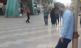 A man wearing a light blue shirt is leaning against a pole in a shopping mall. There are several people walking in the background, including a man in a red shirt, a woman in a black abaya, and a group of people casually dressed. The floor is tiled with a wavy pattern, and the environment includes large green structures and several shops.