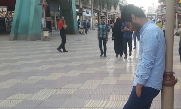 A man wearing a light blue shirt is leaning against a pole in a shopping mall. There are several people walking in the background, including a man in a red shirt, a woman in a black abaya, and a group of people casually dressed. The floor is tiled with a wavy pattern, and the environment includes large green structures and several shops.