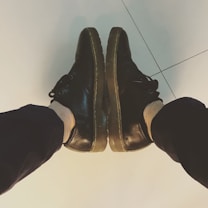 Black leather shoes with thick soles worn by a person sitting with legs extended forward, viewed from above. The shoes are resting on a light-colored tiled floor.
