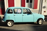 A vintage light blue car is parked on a street next to a building with red shutters. A for-sale sign is visible in the window, indicating the car is being sold for 2500 euros. Sunlight creates a strong shadow on the pavement.