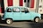 A vintage light blue car is parked on a street next to a building with red shutters. A for-sale sign is visible in the window, indicating the car is being sold for 2500 euros. Sunlight creates a strong shadow on the pavement.