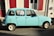 A vintage light blue car is parked on a street next to a building with red shutters. A for-sale sign is visible in the window, indicating the car is being sold for 2500 euros. Sunlight creates a strong shadow on the pavement.