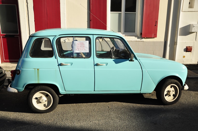 A vintage light blue car is parked on a street next to a building with red shutters. A for-sale sign is visible in the window, indicating the car is being sold for 2500 euros. Sunlight creates a strong shadow on the pavement.