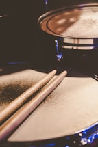 A close-up of drumsticks resting on a snare drum, bathed in warm stage lighting.