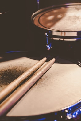 A close-up shot of a drum set, focusing on the drumsticks resting on a snare drum. The lighting creates a warm and slightly moody ambience, with a soft reflection on the drum surface.