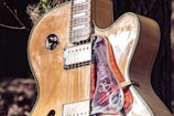 A close-up of a glossy electric guitar featuring a natural wood finish and shiny metal components. The guitar is leaned against a tree, which can be seen in the background. Its pickguard has a distinctive design that includes white scribbles or an autograph.